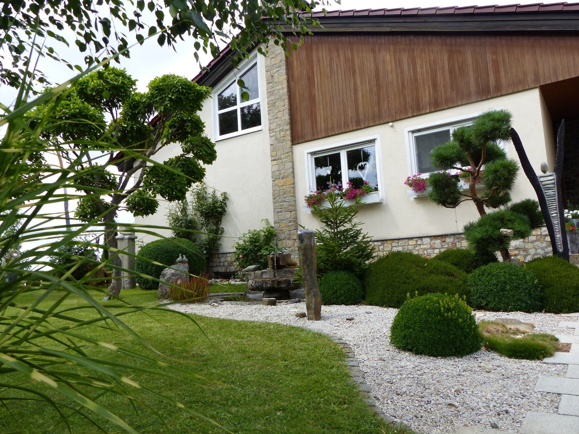 Modern gestalteter Vorgarten mit asiatischem Flair, kunstvoll geschnittenen Bäumen und Kiesflächen. Ein gepflegtes Einfamilienhaus mit Natursteinfassade, Fensterblumen und stilvoller Bepflanzung. Harmonische Gartenlandschaft mit Zen-Elementen.