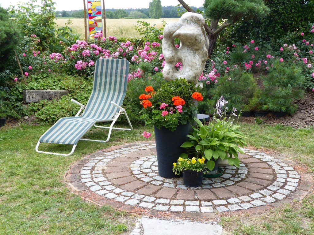 Gartenidylle mit Relaxliege, moderner Steinskulptur und blühenden Blumen. Ein gemütlicher Sitzbereich mit gepflastertem Rundplatz, umgeben von grünen Pflanzen und pinken Rosensträuchern. Perfekter Ort zur Entspannung im Freien